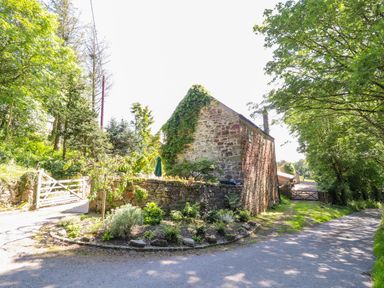 A stone building with ivy on the wall surrounded by trees and two gates on a country road at The Old Coach House in Llanarmon-Yn-Ial