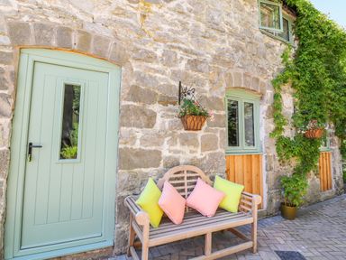 A stone wall with a light green door and windows a wooden bench with colorful cushions and hanging flower baskets at The Old Coach House in Llanarmon Yn Ial