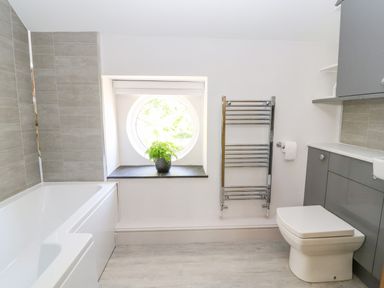 A bathroom with a bathtub a circular window with a plant a towel radiator and a toilet with grey cabinets at The Old Coach House in Llanarmon Yn Ial