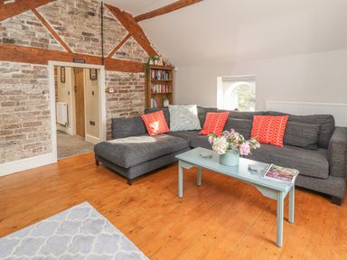 A living room with a gray sectional sofa with red and patterned cushions a light blue coffee table with flowers and magazines a bookshelf and exposed brick walls at The Old Coach House in Llanarmon Yn Ial
