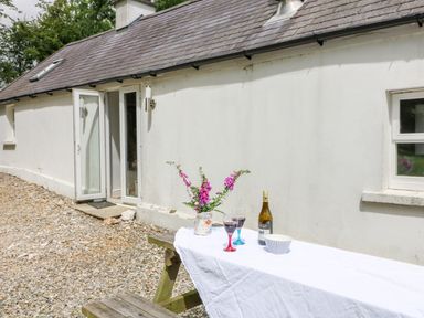 An outdoor area with a wooden picnic table covered with a white cloth holding a vase with flowers two glasses of wine a bottle of wine and a bowl outside a white building with a door and window at The Old White Cottage in Shillelagh County Wicklow