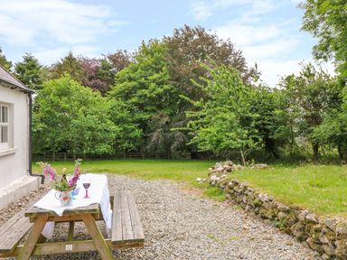 A wooden picnic table with a white tablecloth and flowers on a gravel area next to a white building surrounded by trees at The Old White Cottage in Shillelagh County Wicklow