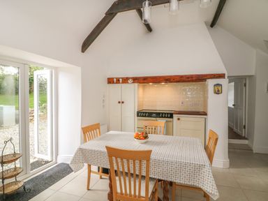 A dining area with a table covered with a patterned tablecloth and four wooden chairs next to a kitchen space with a stove and cupboard at The Old White Cottage in Shillelagh County Wicklow