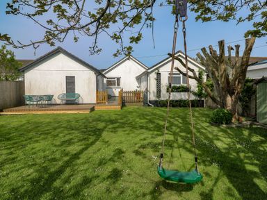 A backyard with a green swing hanging from a tree a wooden deck with green outdoor furniture and white buildings in the background at Tremar Carbis Bay