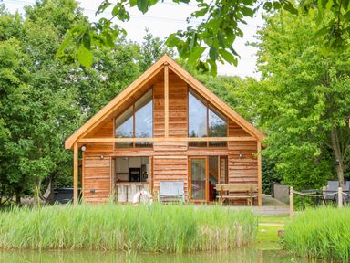 A wooden cabin with large windows and an open door facing a pond surrounded by grass and trees at Dorado Lodge in Thorpe-on-the-Hill