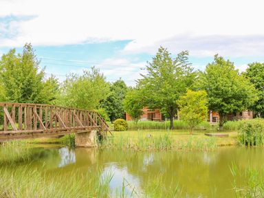 A wooden footbridge over a pond with trees and grass around at Dorado Lodge in Thorpe-on-the-Hill