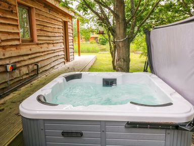 An outdoor hot tub next to a wooden cabin with a tree and grass visible at Dorado Lodge in Thorpe-on-the-Hill