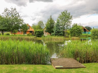 A pond with reeds and trees around it and wooden huts in the background at Dorado Lodge in Thorpe-on-the-Hill