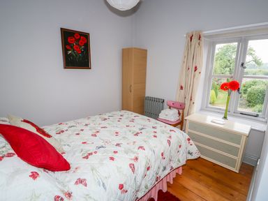A bedroom with a bed with floral bedding a wooden wardrobe a chair with folded towels a chest of drawers and a window with floral curtains at Southrepps Lodge in Southrepps