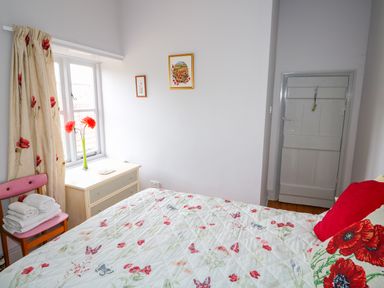 A bedroom with a bed covered in a floral and butterfly patterned quilt next to a window with floral curtains and a small dresser with flowers at Southrepps Lodge in Southrepps