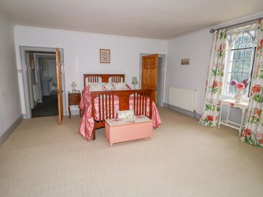 A bedroom with a wooden bed covered with pink bedding a pink storage chest floral curtains and a window at Southrepps Lodge in Southrepps
