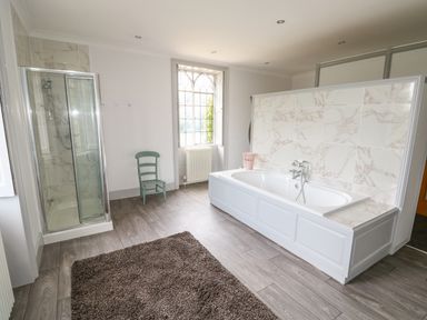 A bathroom with a bathtub surrounded by marble tiles a glass shower enclosure a green chair a window and a brown rug on the floor at Southrepps Lodge in Southrepps