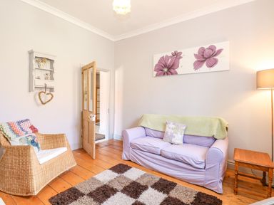 A living room with a purple sofa a wicker armchair a wooden side table a floor lamp and floral wall art at Southrepps Lodge in Southrepps