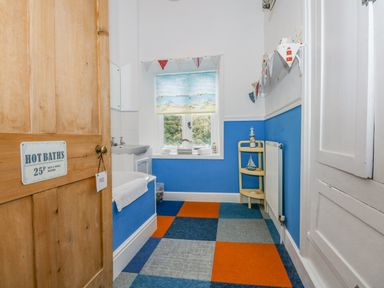 A bathroom with blue and orange checkered carpet a bathtub with a towel a small sink a window with a seaside-themed blind and a wooden door with a hot baths sign at Southrepps Lodge in Southrepps