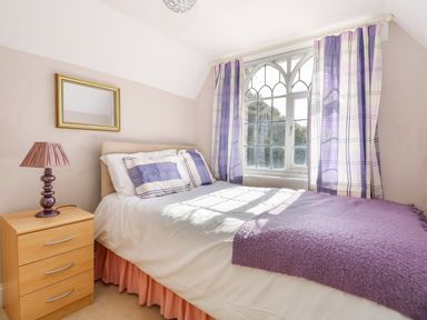 A bedroom with a bed a nightstand lamp and arched window with purple curtains at Southrepps Lodge in Southrepps