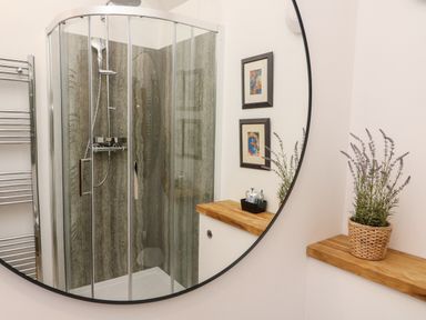 A bathroom with a glass shower enclosure a round mirror two framed pictures and a wooden shelf with a potted plant at Sunshine Cottage in Preston under Scar near Leyburn