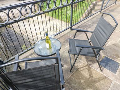 A patio with two black chairs and a glass table holding a bottle of wine and two wine glasses at Sunshine Cottage in Preston under Scar near Leyburn