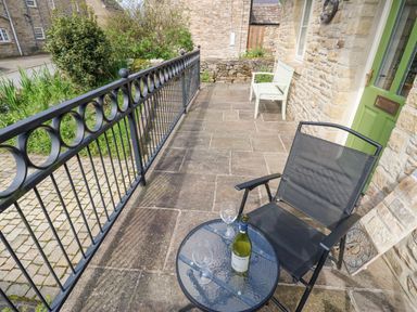 A patio with a black chair and glass table holding a bottle and two glasses outside a stone building at Sunshine Cottage in Preston under Scar near Leyburn