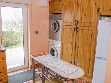 A kitchen with a glass door a washing machine and dryer stacked wooden cabinets a small table with placemats and metal chairs at Ellens Cottage Mount Charles County Donegal