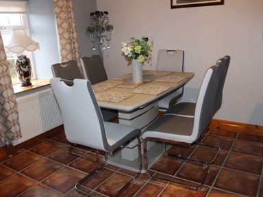 A dining area with a table and six chairs a vase with flowers on the table a lamp on the windowsill and curtains at Ellens Cottage Mount Charles County Donegal
