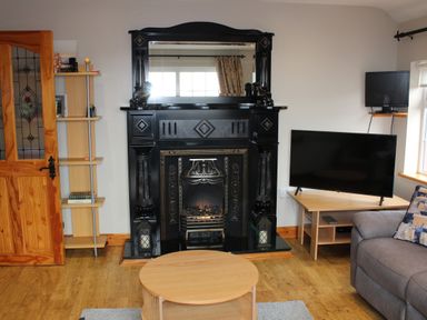 A living room with a black fireplace a wooden door a wooden shelf a TV on a stand a gray couch and a round wooden coffee table at Ellens Cottage in Mount Charles County Donegal