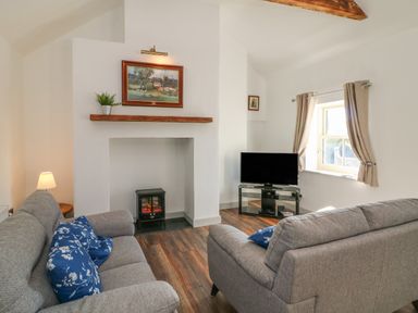 A living room with grey sofas and blue floral cushions a fireplace a TV on a stand and a window with curtains at Macreddin Rock Holiday Cottage Macreddin Aughrim County Wicklow