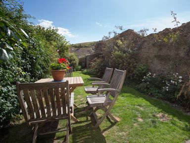 A garden with wooden chairs and a table with a flower pot surrounded by plants and a stone wall at Brock Cottage in West Bay
