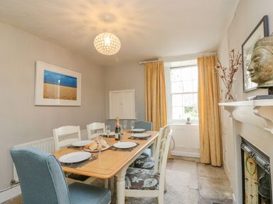 A dining room with a wooden table set for six near a window with yellow curtains at Brock Cottage in West Bay