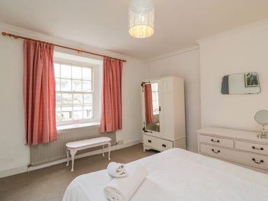 A bedroom with a bed folded towels on it a white dresser a mirror a white wardrobe a bench under a window with pink curtains at Brock Cottage in West Bay