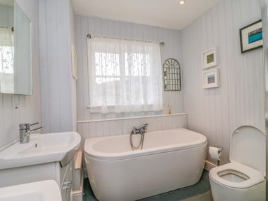A bathroom with a bathtub, sink, toilet, window with sheer curtains, and framed pictures at Brock Cottage in West Bay