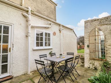 An outdoor patio with a metal table and six chairs next to a white wall with windows at Brock Cottage in West Bay