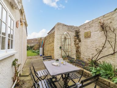An outdoor patio with a metal table and six chairs near a building with garden plants and stone walls at Brock Cottage in West Bay