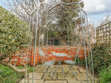 A metal garden table and two chairs under a metal pergola on a stone patio with a brick wall and plants at Brock Cottage in West Bay