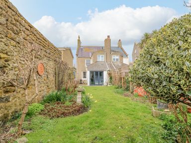 A backyard garden with grass and plants between a stone wall and a house at Brock Cottage in West Bay