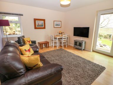 A living room with a brown leather sofa, armchair, small dining table with chairs, wall-mounted TV, and a rug at Plas Marchog Annexe in Rhoscefnhir near Pentraeth