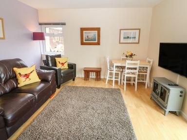 A living room with brown leather sofa and armchair with yellow bird cushions a small dining table with chairs a gray rug and a wall-mounted television at Plas Marchog Annexe in Rhoscefnhir near Pentraeth