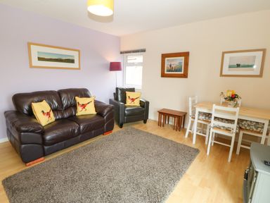 A living room with a brown leather sofa and armchair with bird cushions a dining table with four chairs and framed pictures on the wall at Plas Marchog Annexe in Rhoscefnhir near Pentraeth