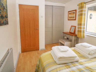 A bedroom with a wooden door, gray closet doors, a small dressing table with mirror, a window with yellow patterned curtains, and a bed with folded white towels at Plas Marchog Annexe in Rhoscefnhir near Pentraeth