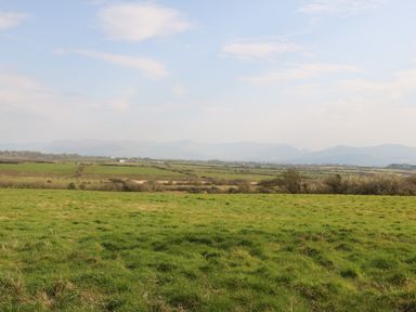 A large green field with trees and distant hills under a partly cloudy sky at Plas Marchog Annexe in Rhoscefnhir near Pentraeth