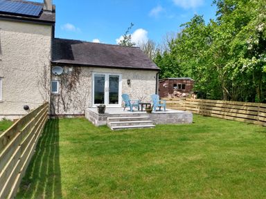 A backyard with grass fenced by wooden panels and a concrete patio with two blue chairs and a table at Plas Marchog Annexe Rhoscefnhir near Pentraeth
