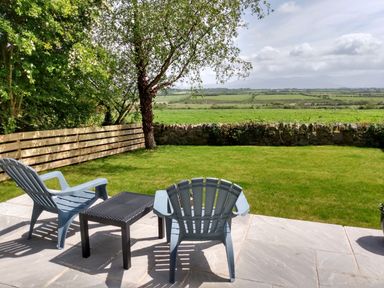 A garden with two chairs and a table on a patio facing green fields with a stone wall and trees at Plas Marchog Annexe in Rhoscefnhir near Pentraeth