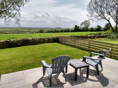 A patio with two chairs and a table overlooking a grassy yard with a low stone wall and a wooden fence at Plas Marchog Annexe in Rhoscefnhir near Pentraeth