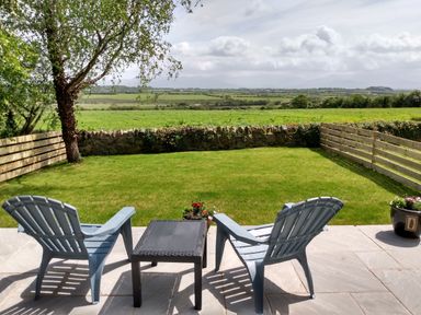 A patio with two chairs and a table overlooking a fenced grassy yard and distant fields at Plas Marchog Annexe in Rhoscefnhir near Pentraeth