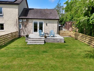 A backyard with a stone patio terrace featuring two blue chairs and a small table surrounded by grass and wooden fencing at Plas Marchog Annexe in Rhoscefnhir near Pentraeth