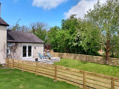 A backyard with a wooden fence grass a small patio with two chairs and a table and trees at Plas Marchog Annexe in Rhoscefnhir near Pentraeth