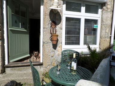 An outdoor patio with green chairs and table with a bottle and glasses and a small dog at the open green door at Labour in Vain in St Ives