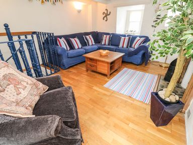 A living room with a blue sectional sofa with striped cushions a wooden coffee table a brown armchair a potted plant and a striped rug at Labour in Vain in St Ives