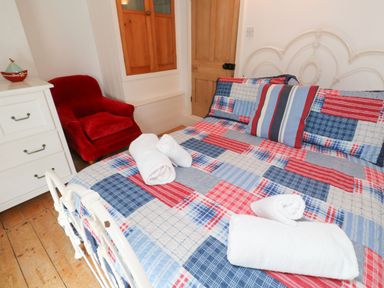 A bedroom with a bed with a patchwork bedspread and rolled white towels a red armchair and a white chest of drawers at Labour in Vain in St Ives
