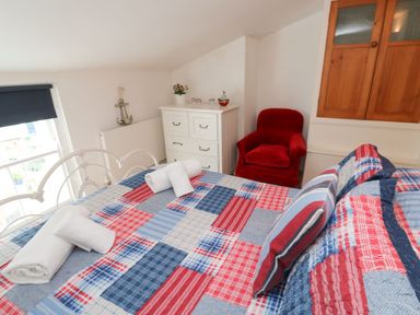 A bedroom with a bed covered in a red blue and white patchwork quilt two rolled white towels on the bed a red armchair a white chest of drawers with a plant and decoration on top and wooden cupboards on the wall at Labour in Vain in St Ives