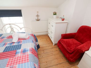 A bedroom with a bed covered in a red blue and white quilt near a white chest of drawers and a red armchair at Labour in Vain in St Ives